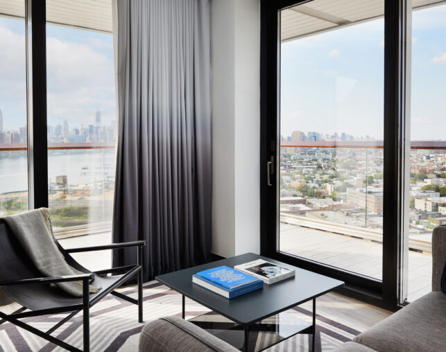 Lounge area with armchair, coffee table, and floor-to-ceiling windows overlooking city skyline.