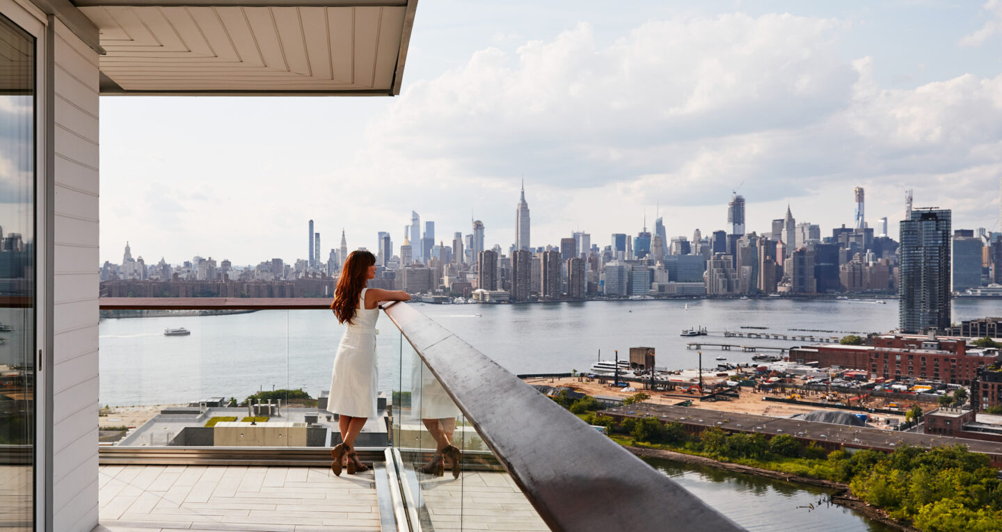Woman in white dress standing on wraparound balcony, admiring Manhattan skyline across the river.