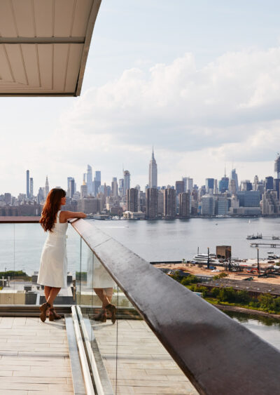 Woman in white dress standing on wraparound balcony, admiring Manhattan skyline across the river.