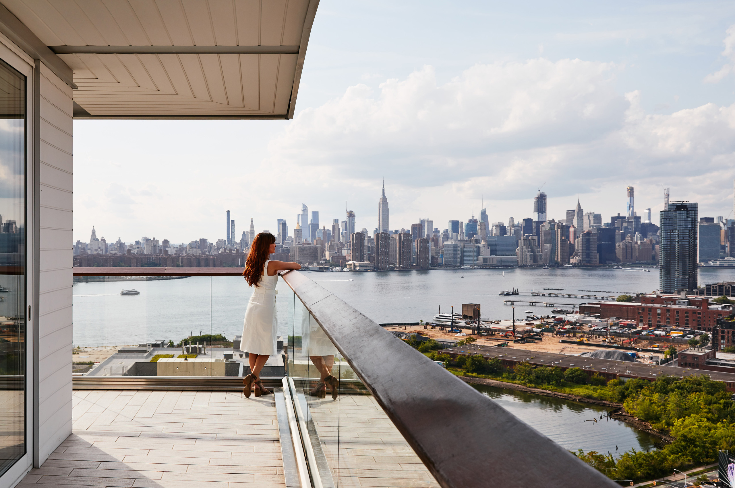 Woman in white dress standing on wraparound balcony, admiring Manhattan skyline across the river.