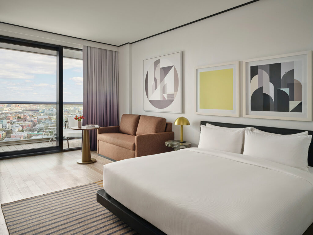 Hotel room with king bed, brown sofa, abstract artwork, and floor-to-ceiling city view