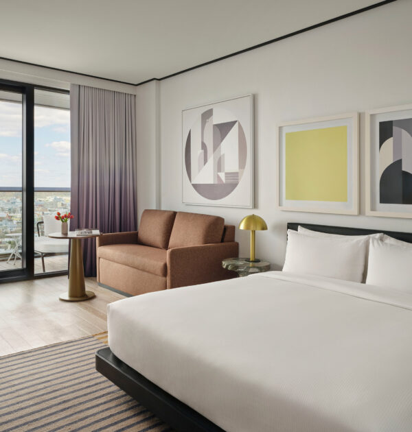 Hotel room with king bed, brown sofa, abstract artwork, and floor-to-ceiling city view