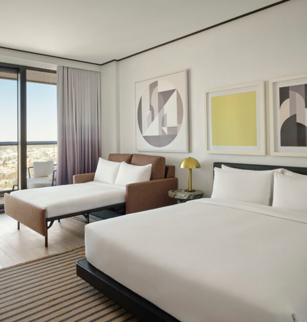 Hotel room with king bed and pull-out sofa bed, large windows overlooking cityscape.