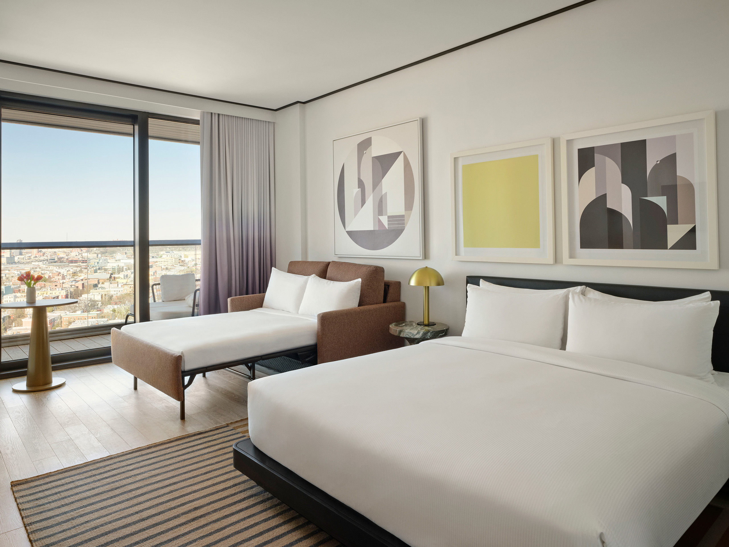 Hotel room with king bed and pull-out sofa bed, large windows overlooking cityscape.