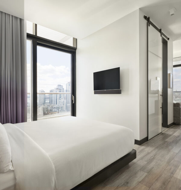Contemporary bedroom with white linens, floor-to-ceiling window, wall-mounted TV, and city view.