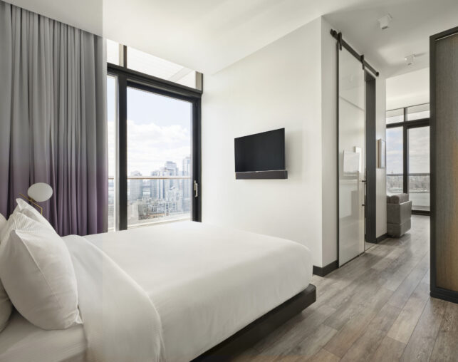 Contemporary bedroom with white linens, floor-to-ceiling window, wall-mounted TV, and city view.