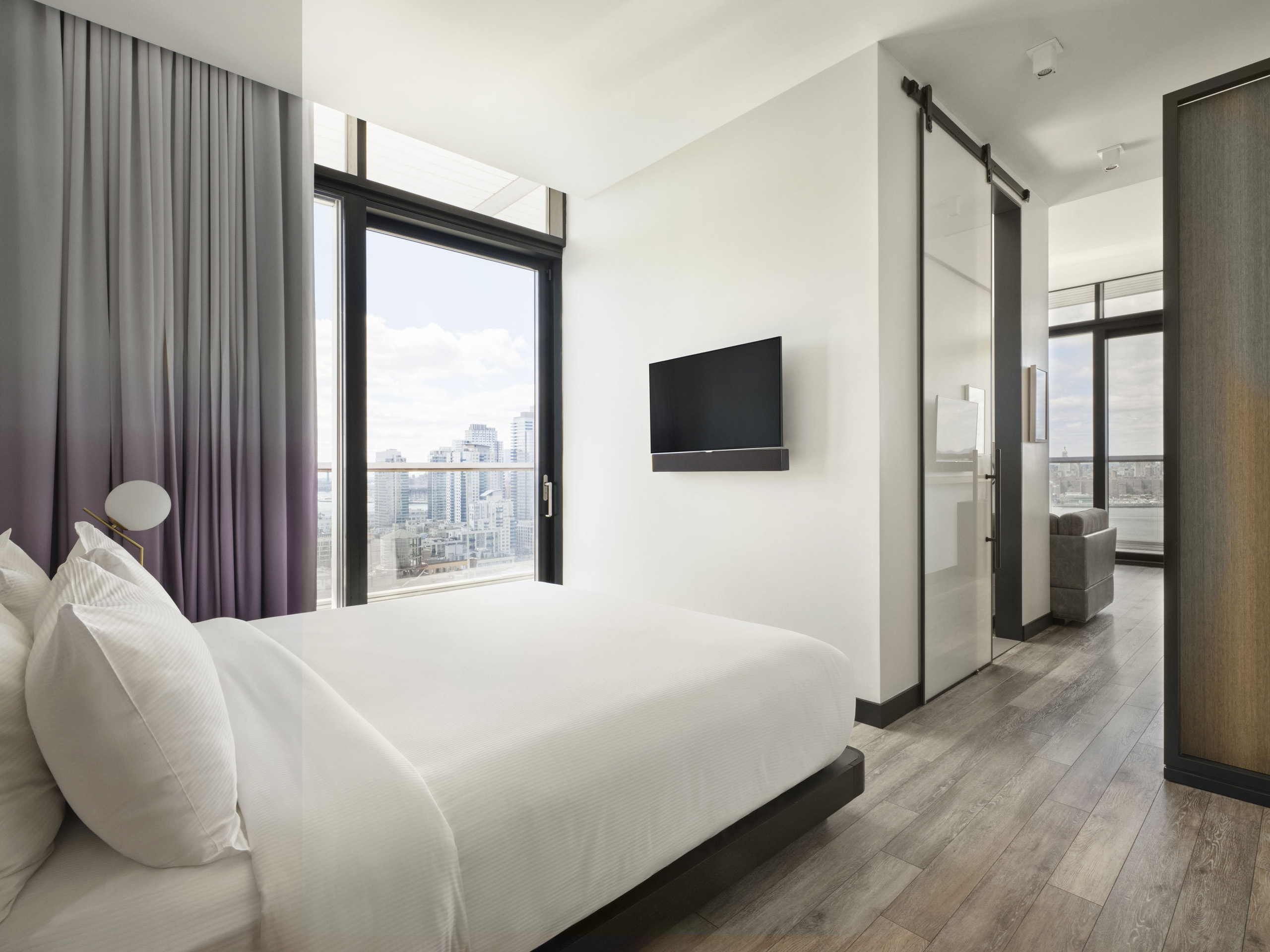 Contemporary bedroom with white linens, floor-to-ceiling window, wall-mounted TV, and city view.