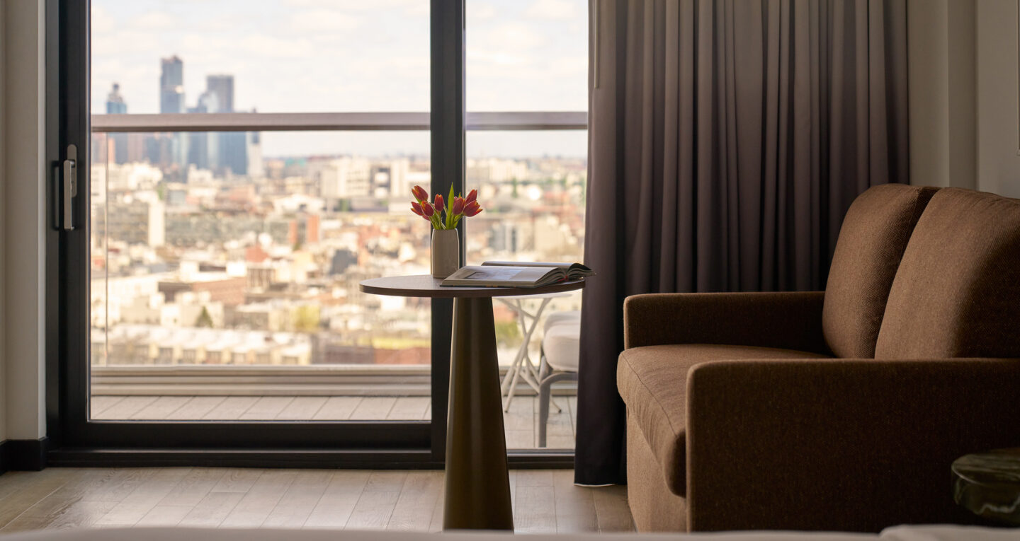 Close-up of brown sofa and round table with tulips beside a window with city view.