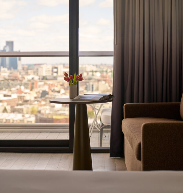 Close-up of brown sofa and round table with tulips beside a window with city view.