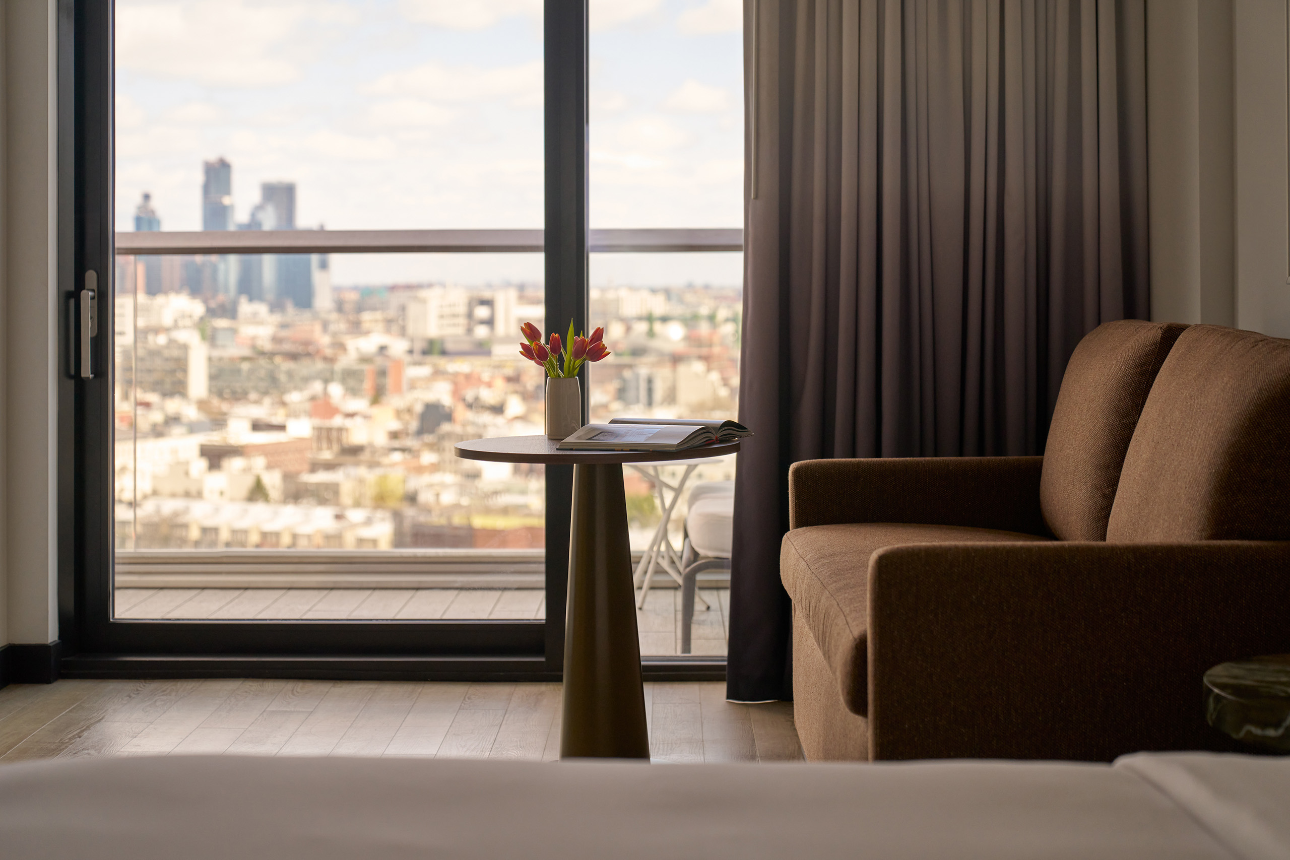 Close-up of brown sofa and round table with tulips beside a window with city view.