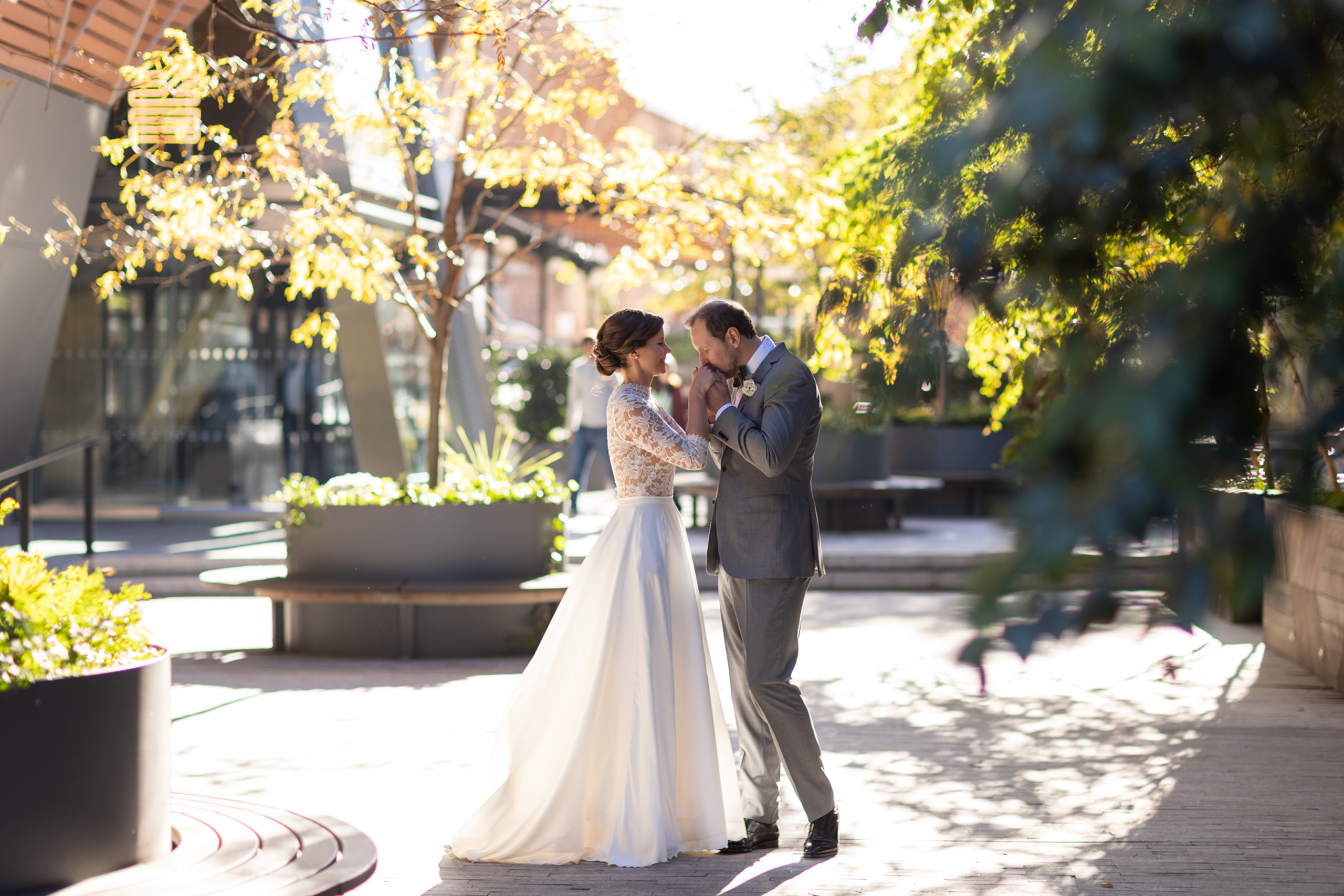 Bride and groom holding hands in a sunlit courtyard surrounded by trees.