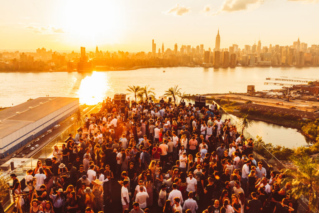 Large rooftop crowd at sunset overlooking New York City skyline.