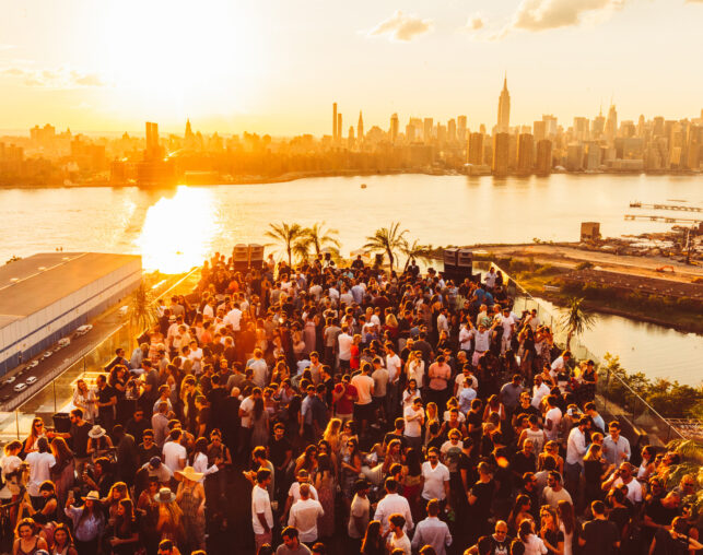 Large rooftop crowd at sunset overlooking New York City skyline.
