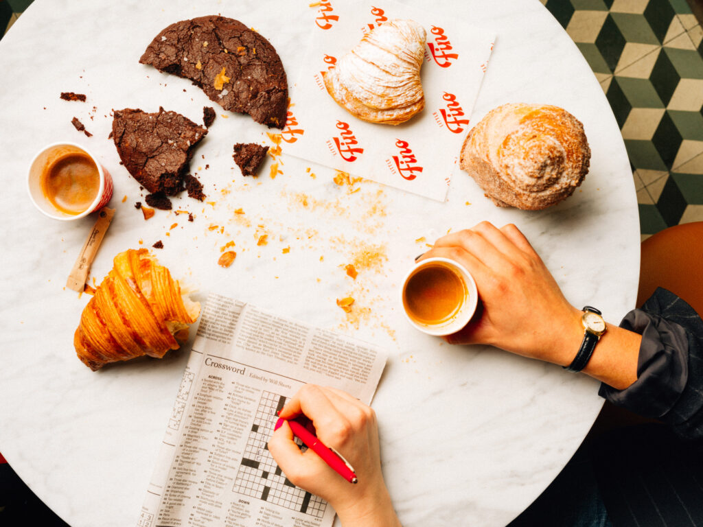Coffee, pastries, and crossword on crumb-covered marble café table.