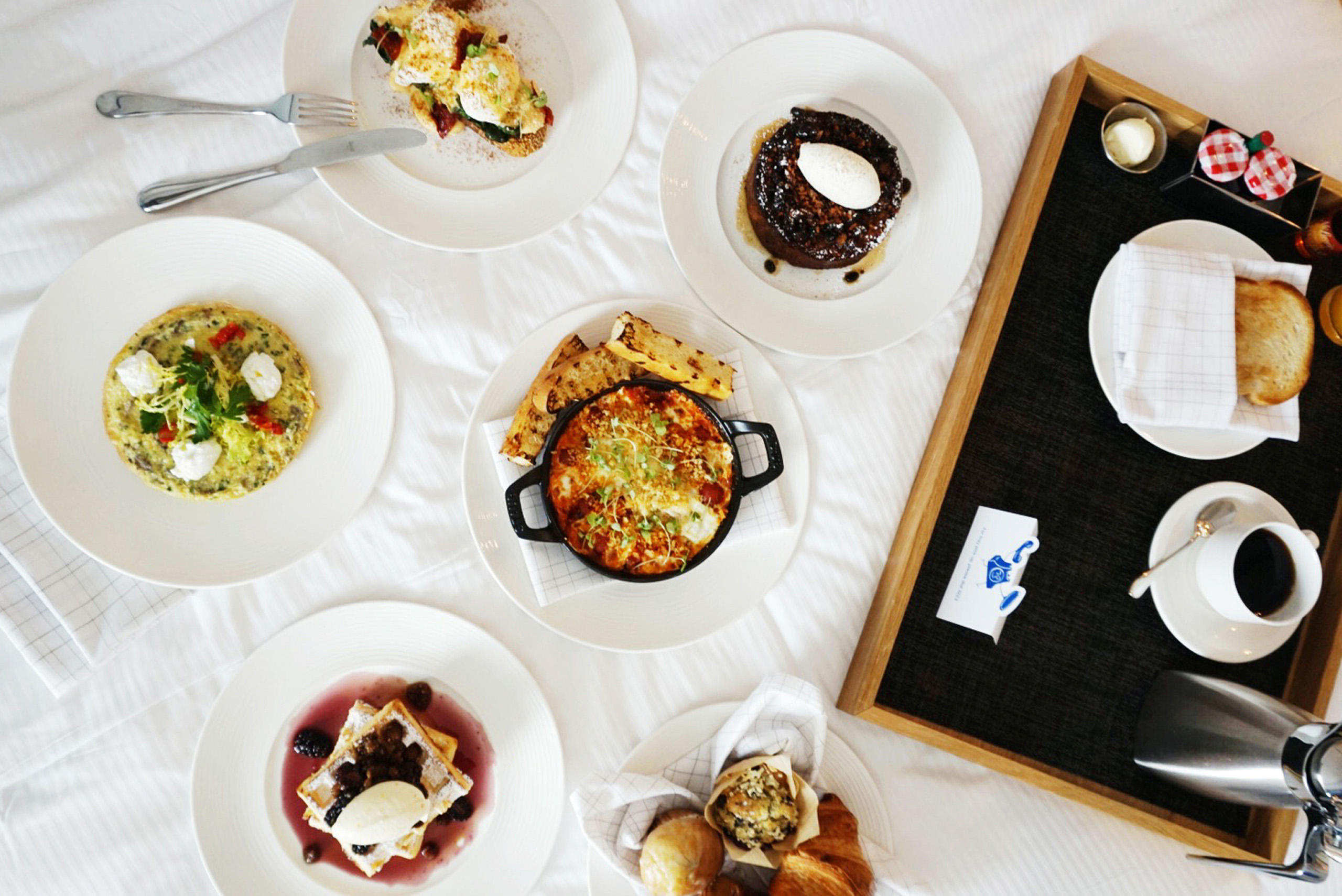 Room service dishes arrayed on a bed and tray, waffles, soup, pancakes, quiche, coffee