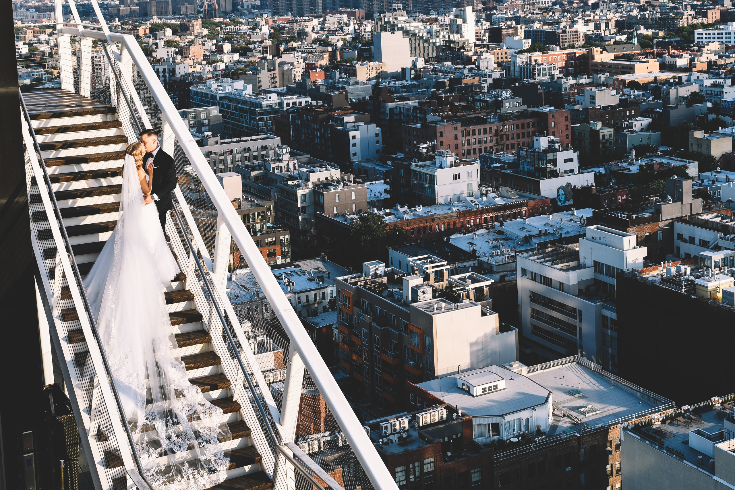 Bride and groom kissing on outdoor stairs overlooking a dense cityscape.