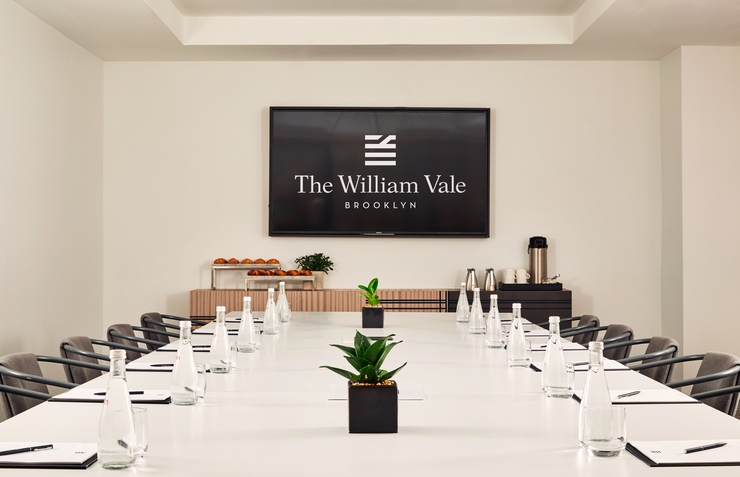 Boardroom with The William Vale screen, notepads, bottled water, and potted plants.