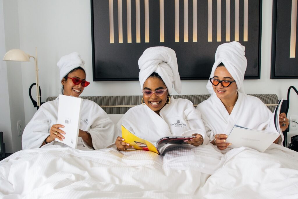 Three women sit in robes in bed laughing and reading magazines.