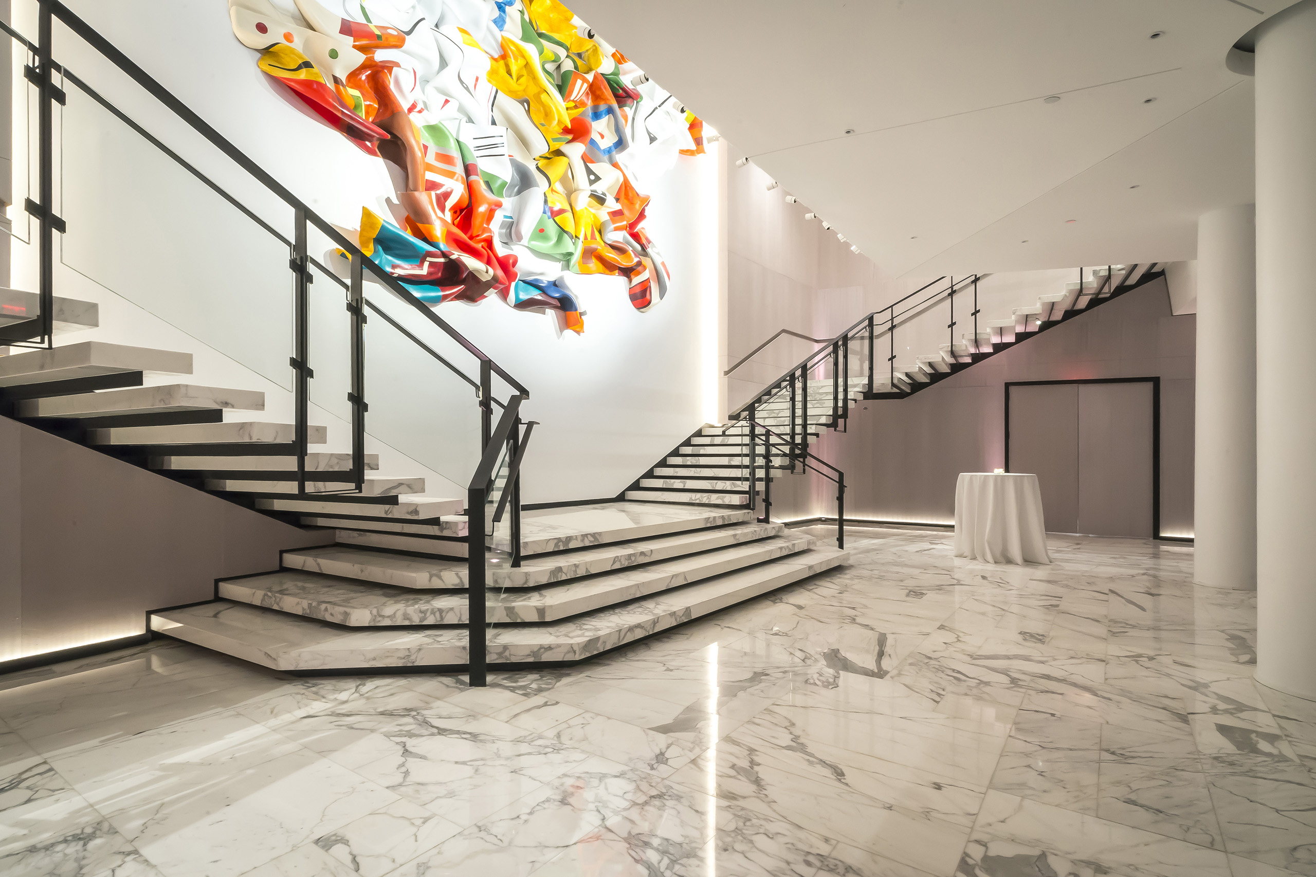 Modern marble staircase with glass railing and colorful abstract wall sculpture.