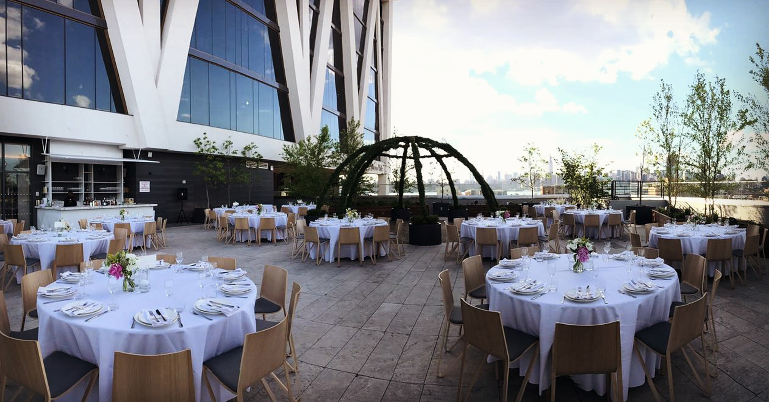 Elegant rooftop reception setup with round tables and floral centerpieces under open sky.