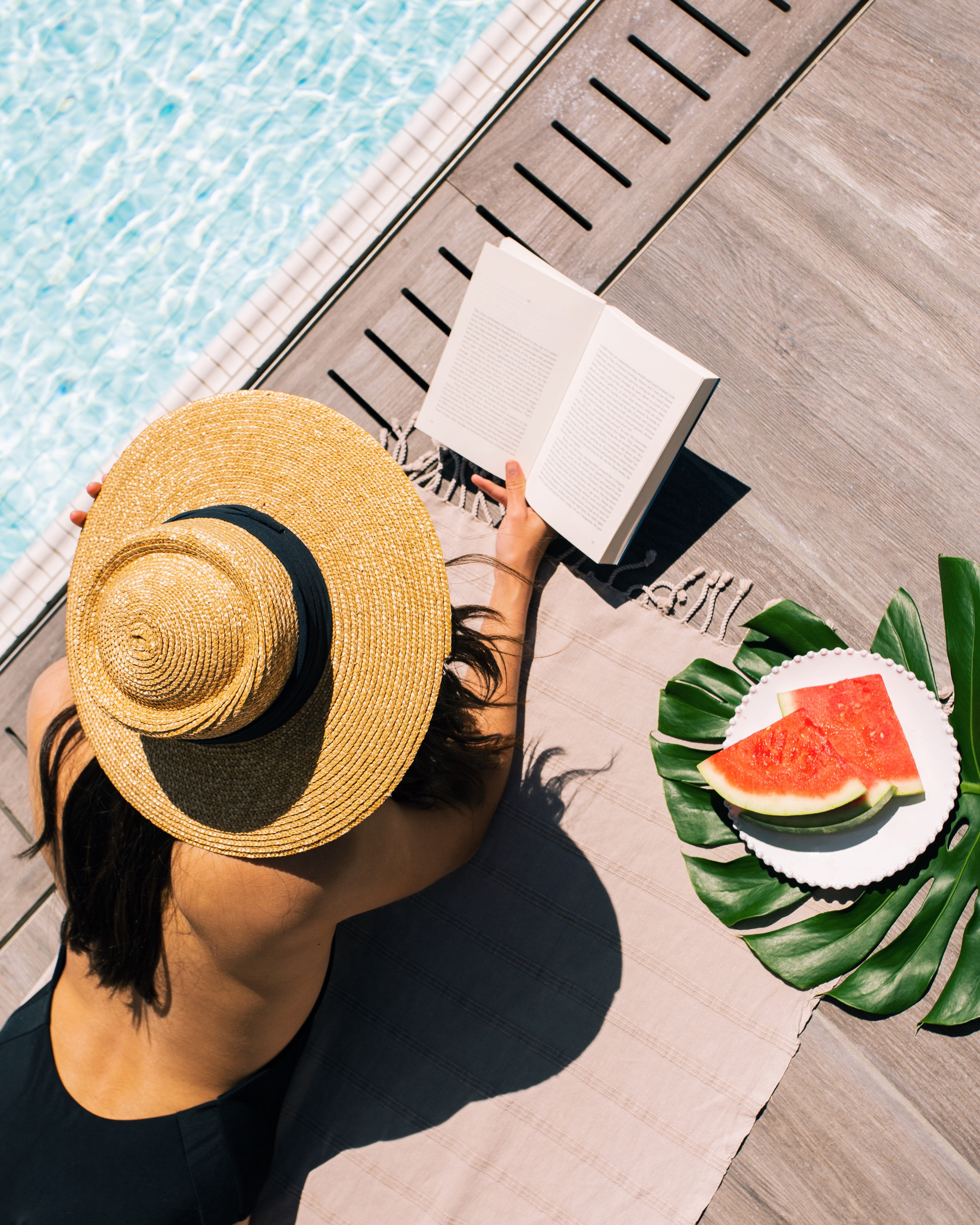 woman in wide brim hat reading a book by the pool with a plate of fresh watermelon
