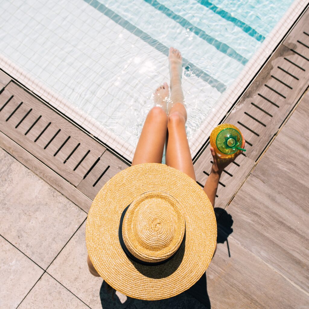 Woman in wide-brim hat relaxing poolside with drink.
