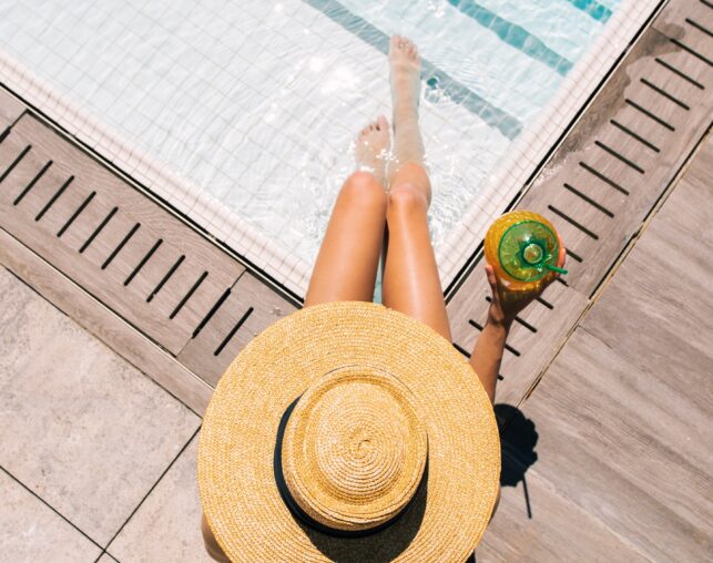 Woman in wide-brim hat relaxing poolside with drink.