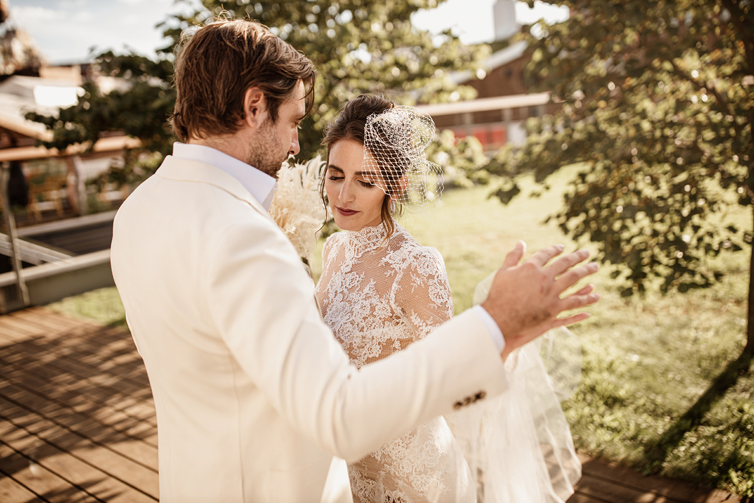 Bride and groom dancing outdoors in sunlight, bride wearing lace dress and birdcage veil.