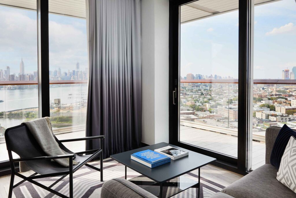 Lounge area with armchair, coffee table, and floor-to-ceiling windows overlooking city skyline.