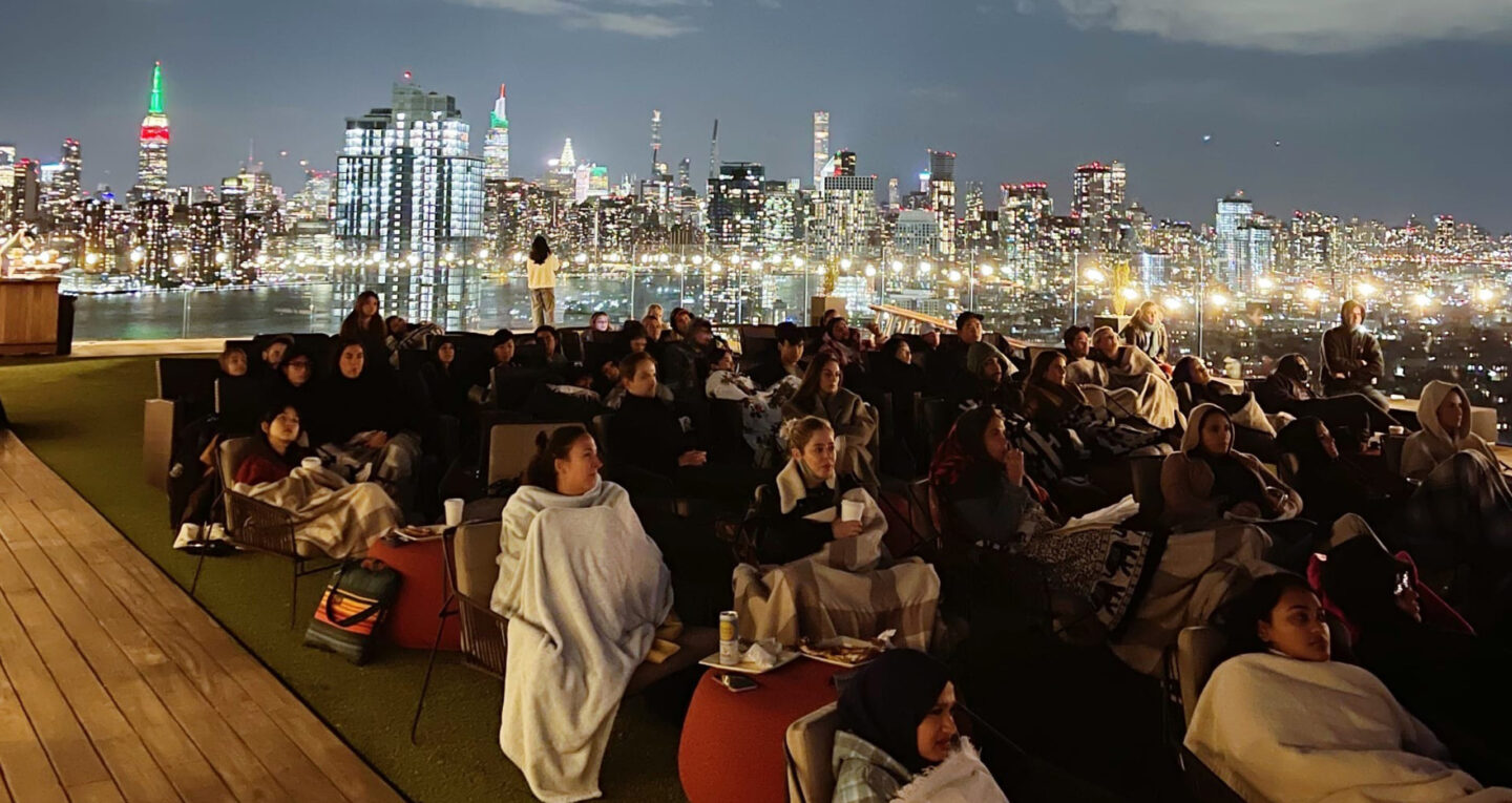 Crowd watching outdoor movie with city skyline at night.