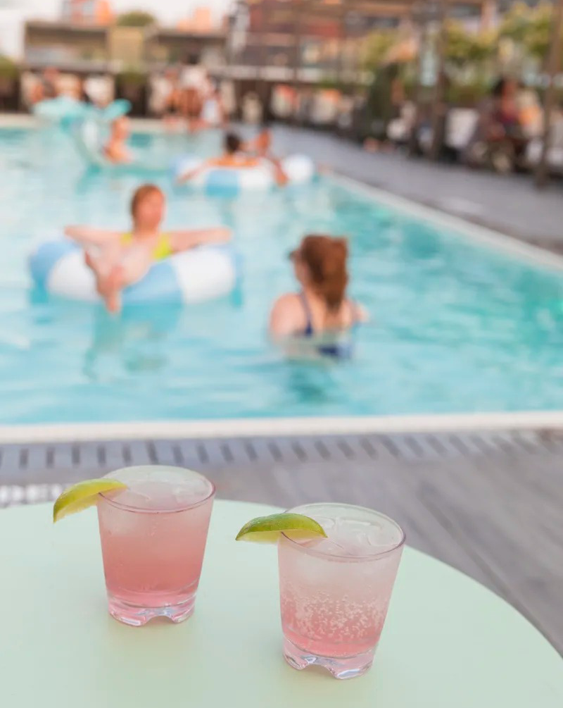 Two pink cocktails by a lively swimming pool.