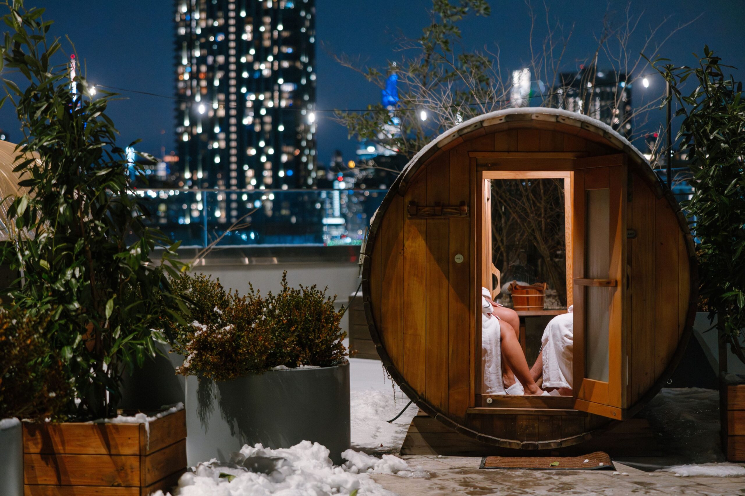 Two guests can be seen sitting inside barrel sauna with Manhattan skyline aglow in the distance.