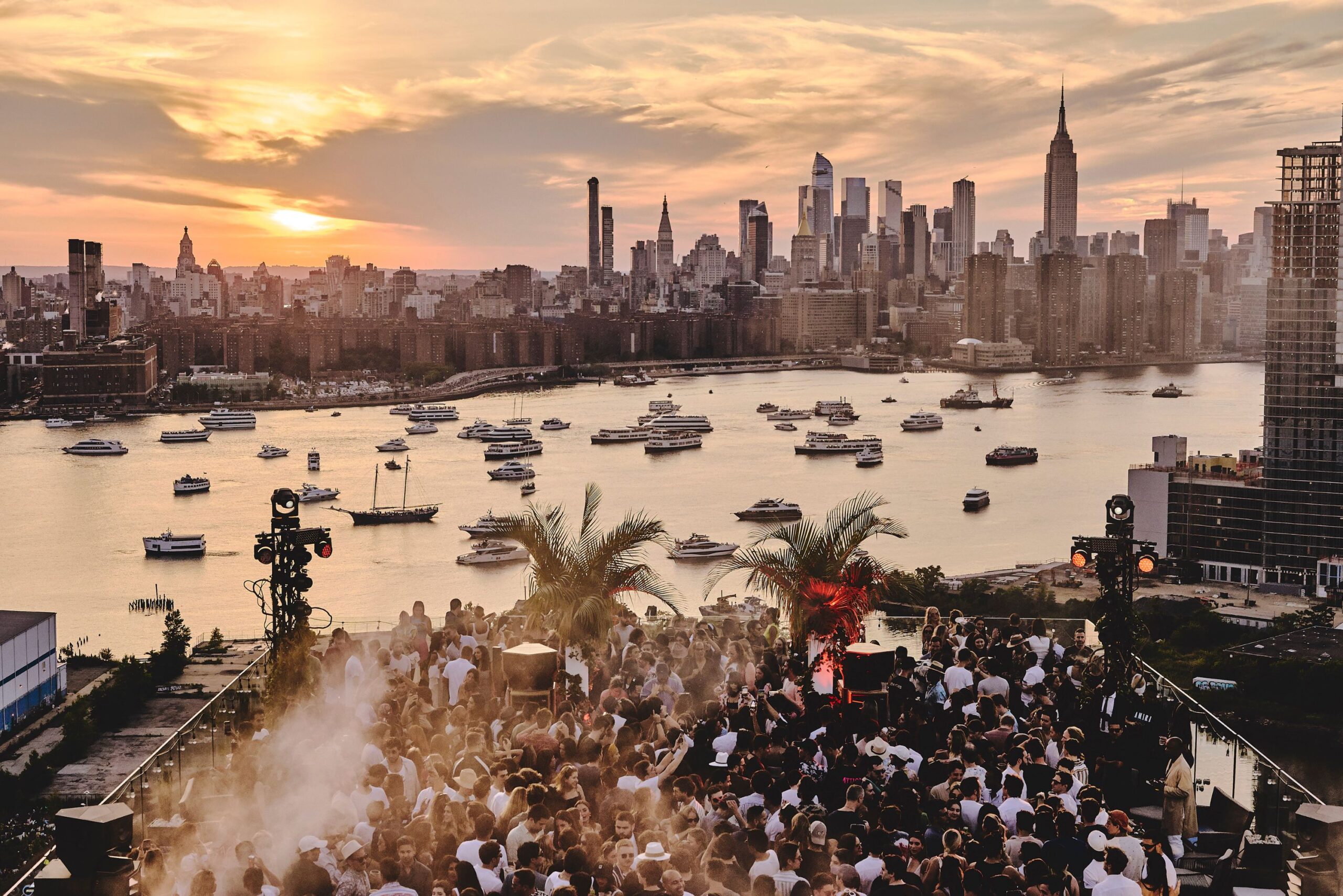 Guests gather on The Turf Club for a party at sunset. The East River and Manhattan skyline are seen in the distance.