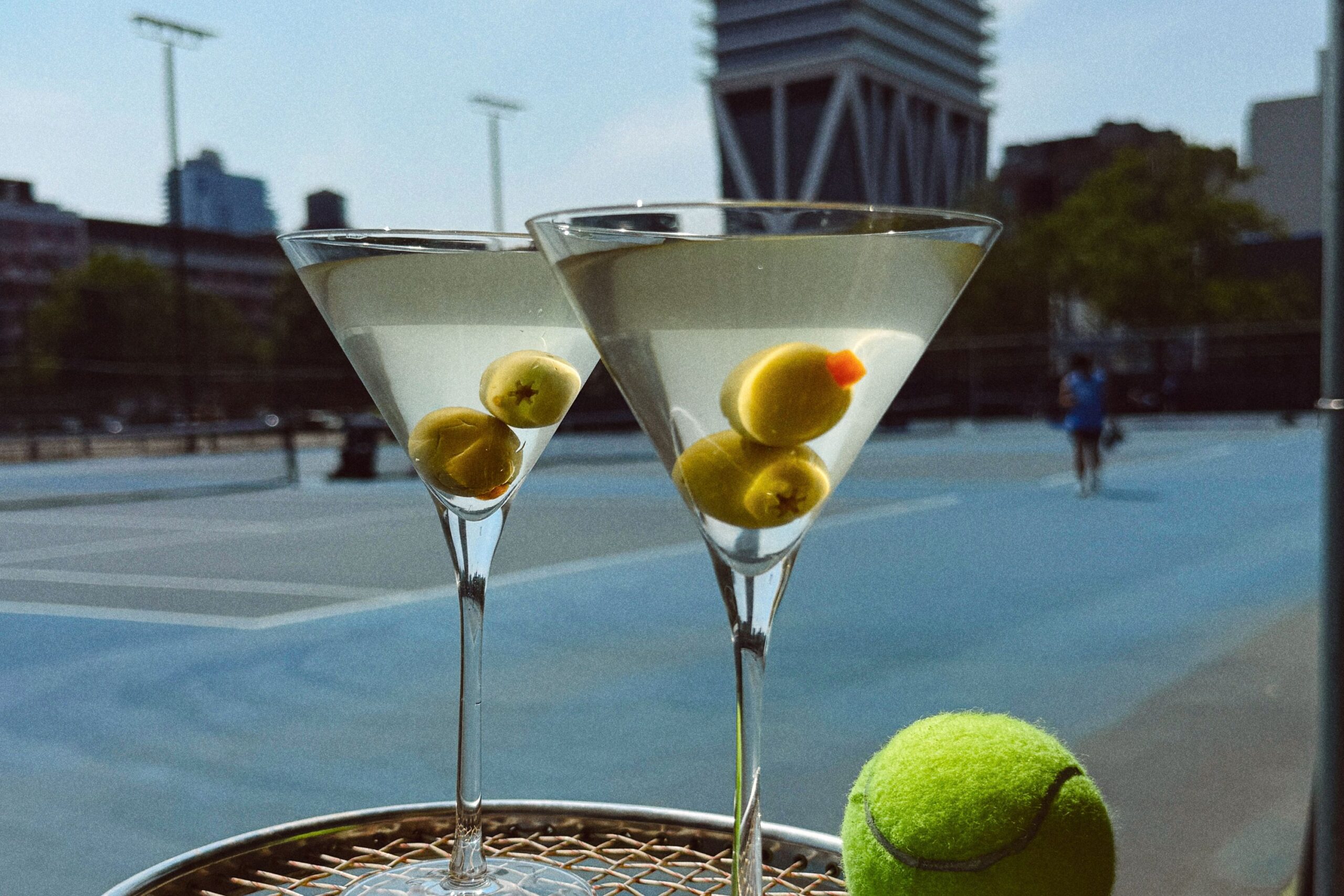 Two martinis sit on a tennis racquet with a tennis ball. The William Vale stands tall in the background.