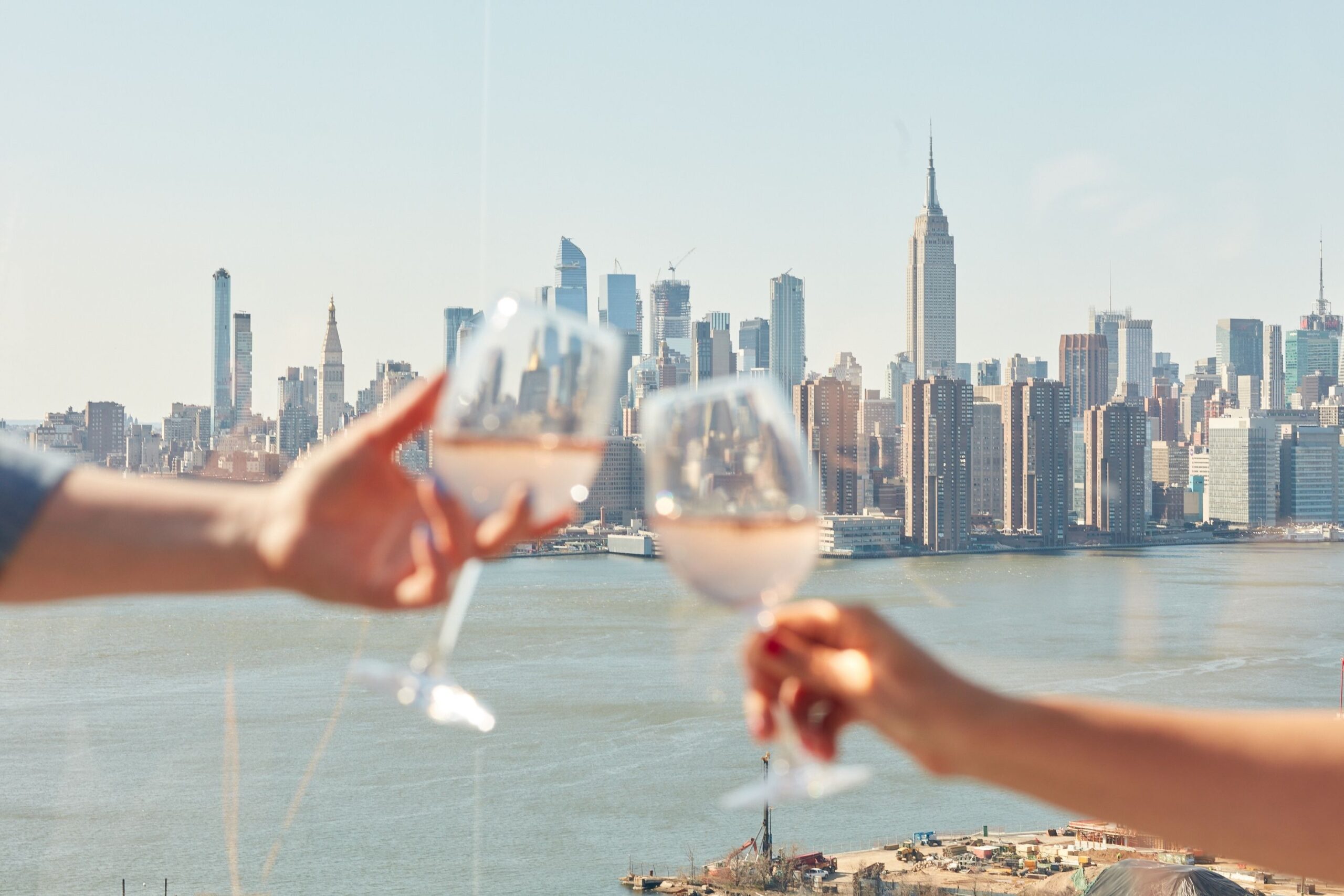 Guests cheers with their glasses from a balcony at The William Vale with the Empire State Building and Manhattan skyline in the distance.