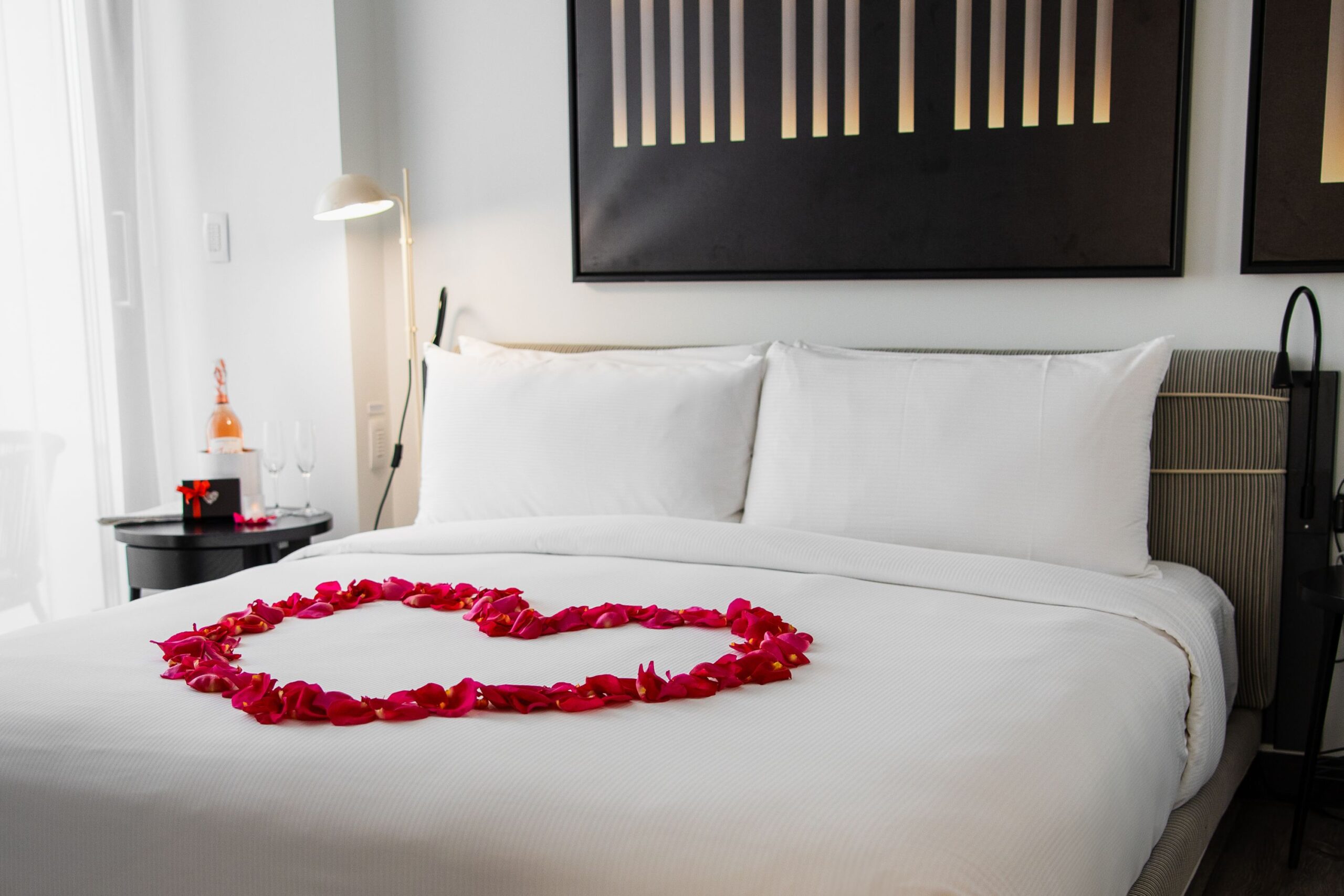 Rose petals are arranged in a heart on the bed. A bottle of wine and box of chocolates sit on the nightstand.