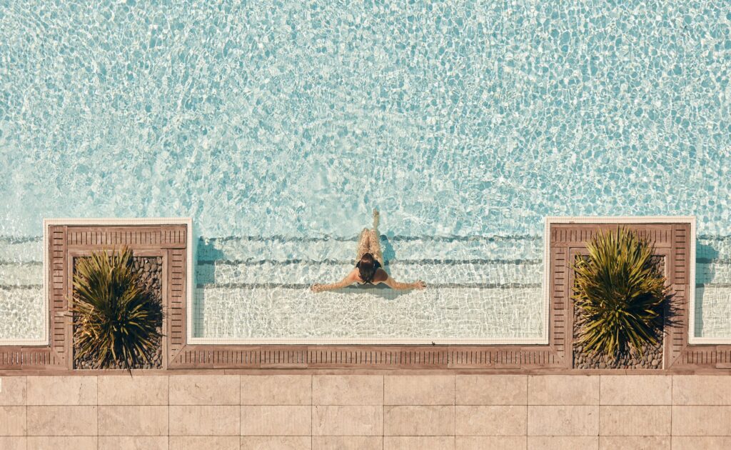 A woman swims in a pool surrounded by beige tiles and palm trees.
