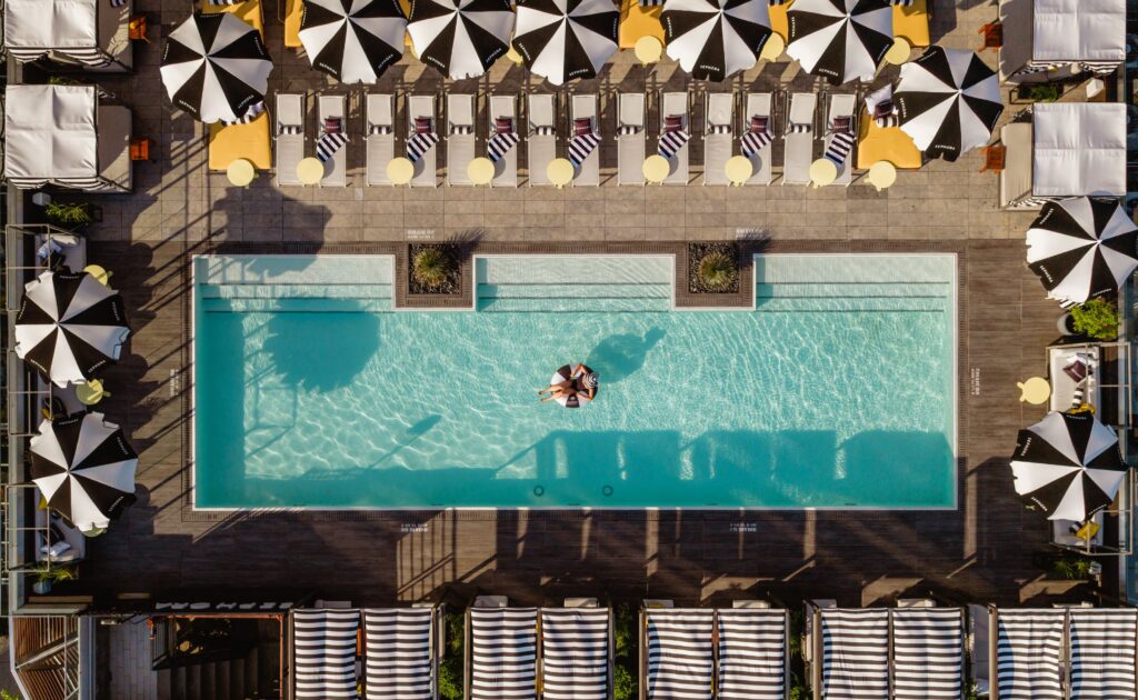 An aerial view of a pool with people in it, surrounded by lounge chairs and umbrellas.