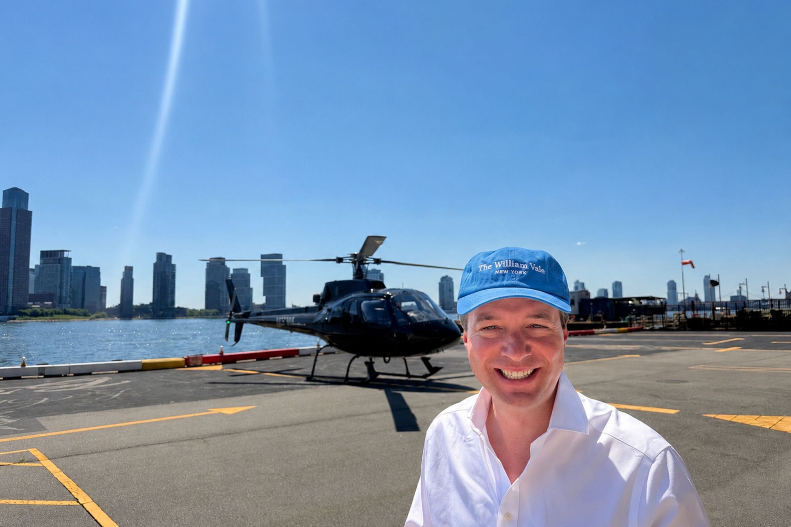 Guest smiles while wearing The William Vale dad hat at helipad with helicopter in the distance.